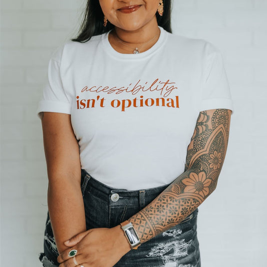 Jenny Jay is pictured wearing a white, tight-fitting t-shirt with her hands clasped in front of her. T-shirt reads 'Accessibility isn't Optional' in orange text.