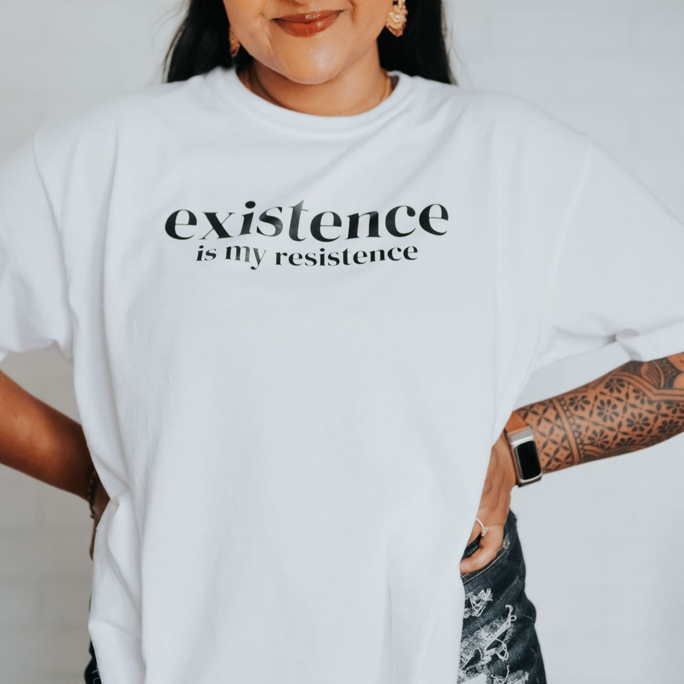 Jenny Jay is pictured wearing a white, oversized t-shirt. T-shirt reads 'Existence is My Resistance' in black text.v