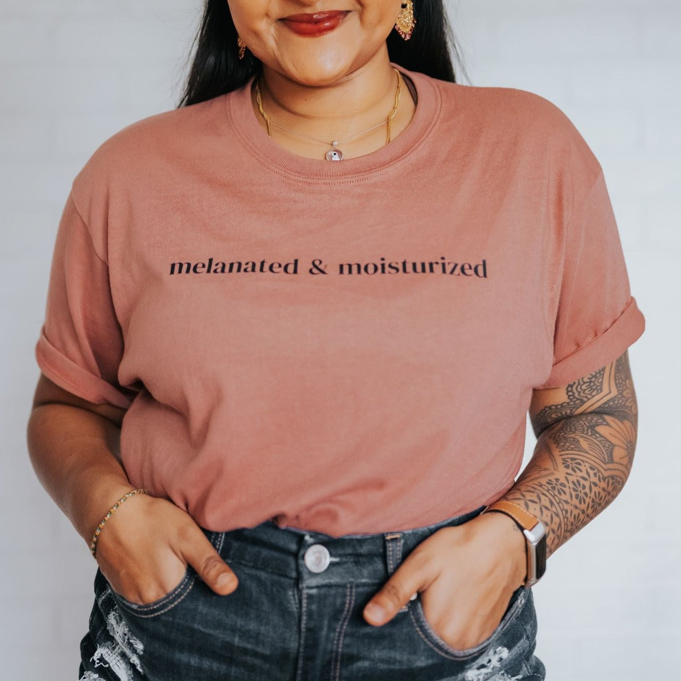 Jenny Jay is pictured wearing a dusty rose t-shirt with her hands in her pockets. T-shirt reads 'Melanated & Moisturized' in black text.